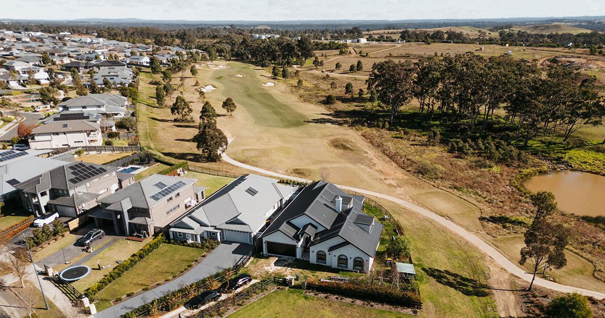 Aerial drone view of a residential estate at Wilton, NSW, showing newly built homes bordering a golf course with fairways, trees, walking paths and surrounding rural landscape under clear skies.