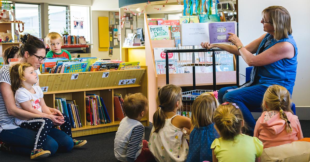 Library staff member reads a picture book to a group of young children seated on the floor during a storytime session in a public library.