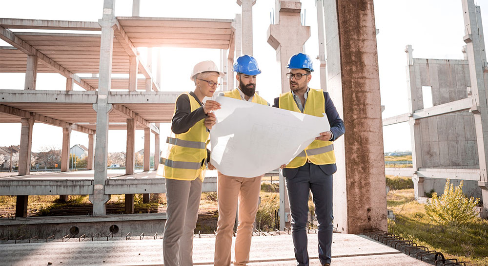 Three people in hard hats look over plans at a construction site.