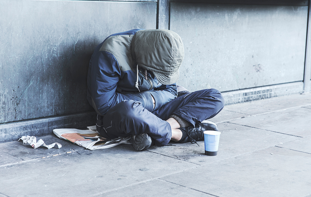 A homeless person sits cross legged on the street wearing a hoodie. 