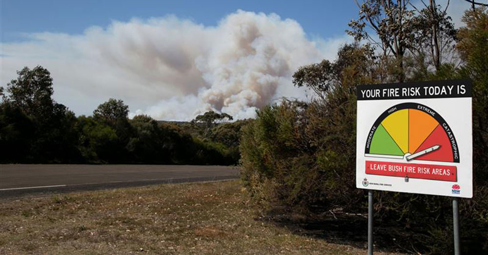 A roadside fire danger rating sign with smoke from a bushfire seen in the distance.