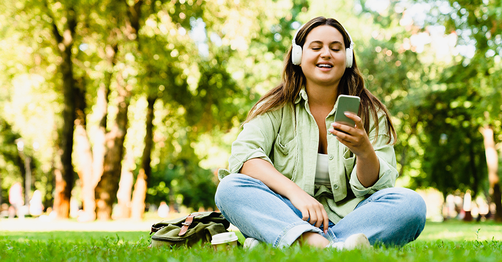 A young woman with headphones reading her smart phone on a grassy spot in a park.