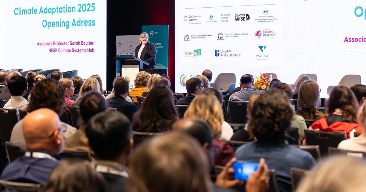 Audience seated at the 2025 Climate Adaptation conference listens to a keynote speaker delivering the opening address from the stage, with sponsor logos displayed on a large screen behind.