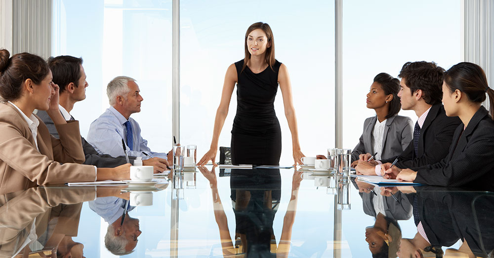 A woman standing at the end of a boardroom table addressing people seated at the table. 