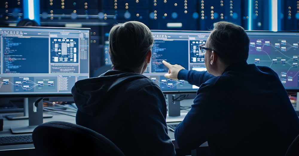 Two IT specialists look at a bank of computer screens with coding.