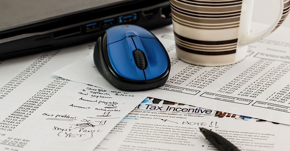 A pen, computer mouse and coffee cup sitting on a bunch of financial papers.