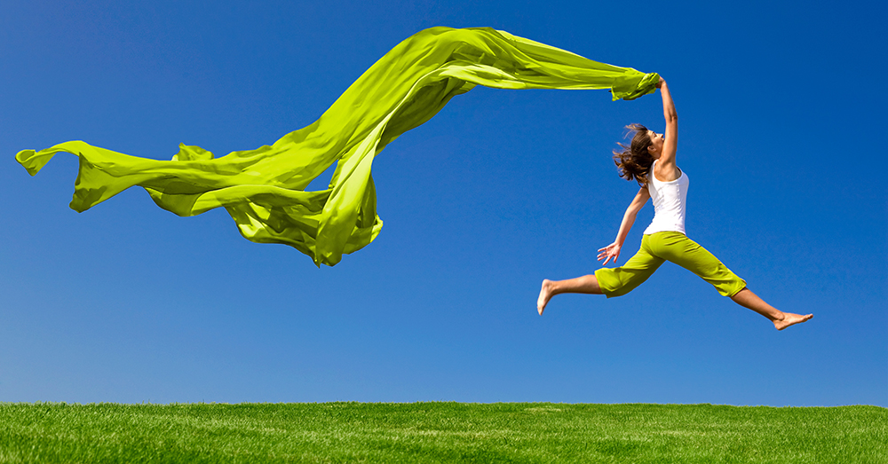 A woman jumping on a bright green field against a bright blue sky with a green ribbon training behind her.