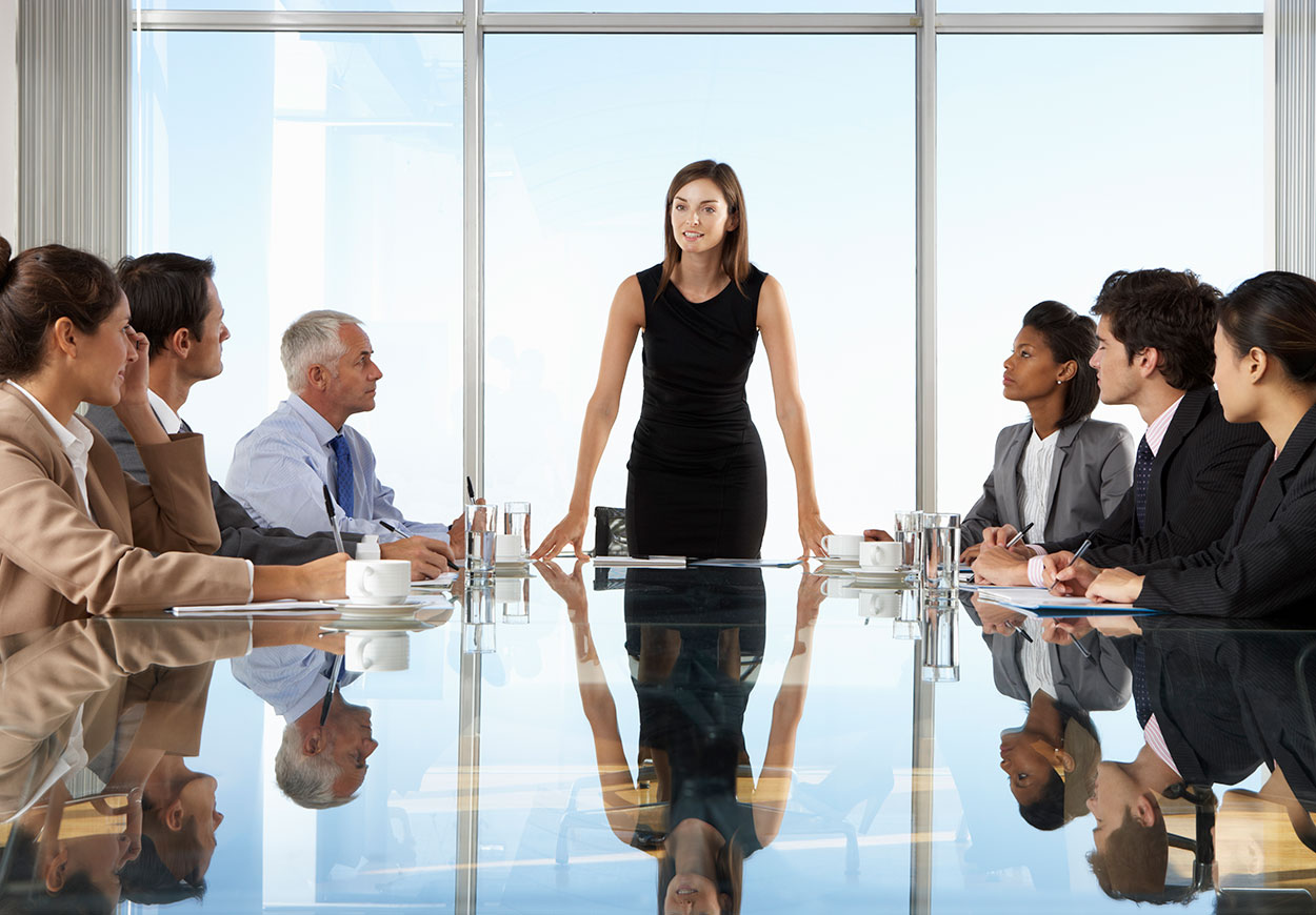 A woman stands at the head of a table to address a meeting.