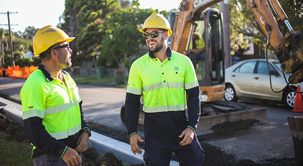 Two road workers in hard hats and high viz vests share a lighter moment.