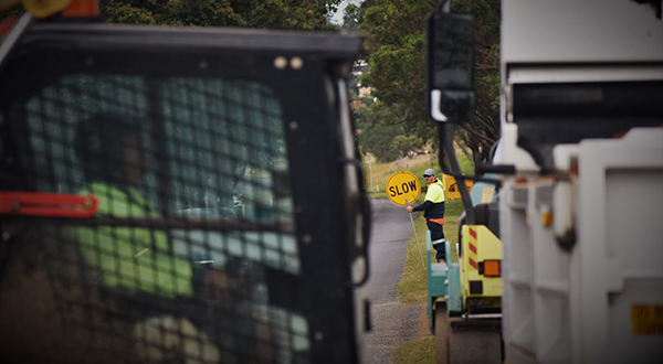 Road worker with a stop/slow sign seen in distance between a truck and Bobcat.