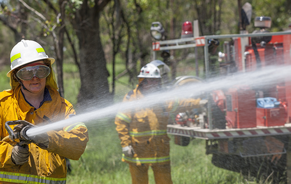 A female rural fire fighter with hose and fire truck. She is dressed in protective clothing.