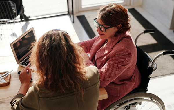 Woman in a wheelchair at desk and computer in discussion with an able-bodied woman.